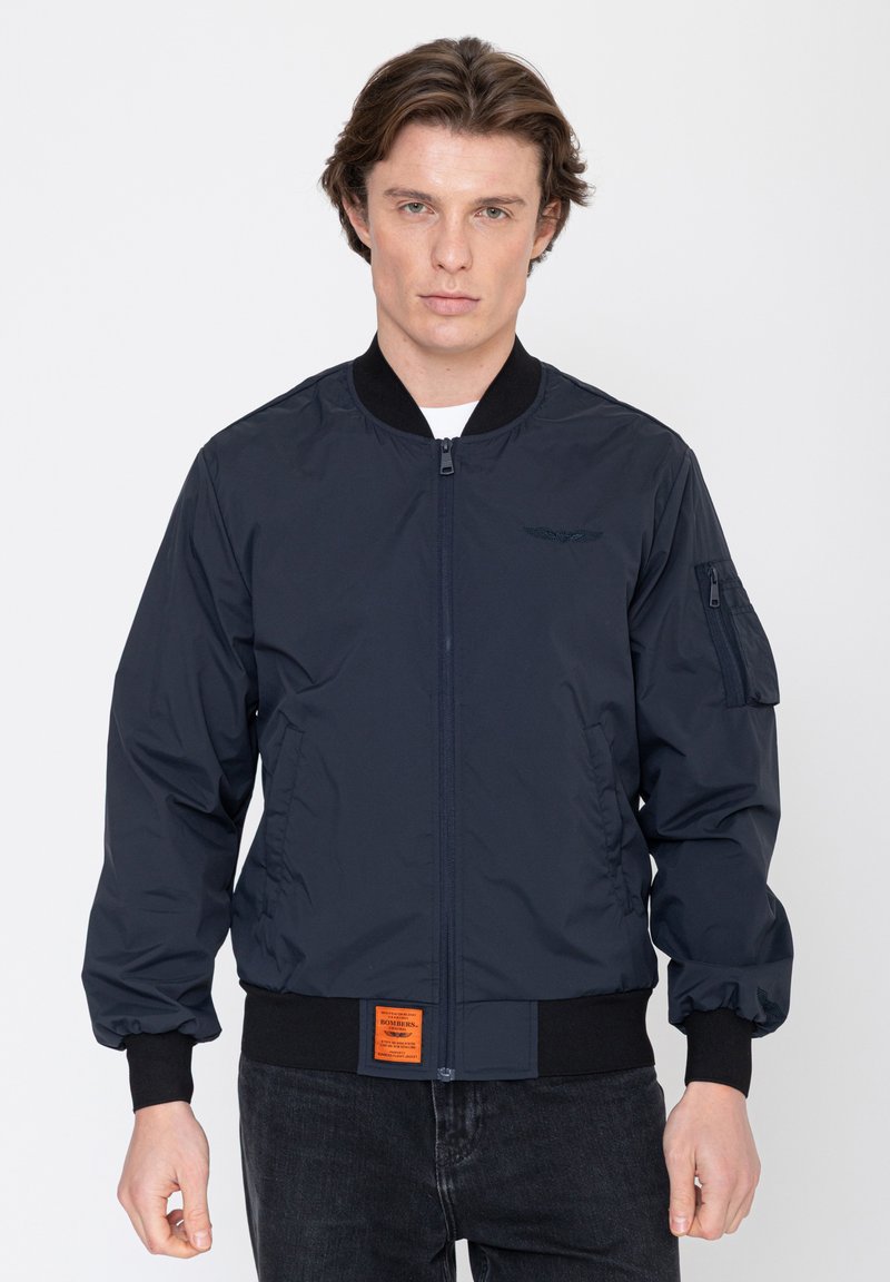 Mann trägt eine marineblaue Bomberjacke mit gerippten Bündchen und Saum, Reißverschluss vorne und einem kleinen orangefarbenen Aufnäher in der Nähe der Taille vor weißem Hintergrund.