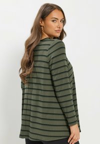 Nainen, jolla on pitkät ruskeat hiukset, pukeutunut ribbitettyyn vihreään pitkähihaiseen puseroon, jossa on vaHorizontal black stripes, katsoo yli olkansa yksinkertaista taustaa vasten.