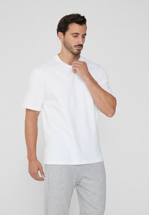 Homme aux cheveux courts et sombres, avec une barbe, portant un t-shirt blanc uni et un pantalon gris clair, se touchant le cou d'une main.