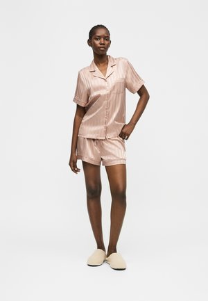 Vrouw staand, draagt lichtroze satijnen kort mouw blouse met knopen en bijpassende korte broek met beige pantoffels op een effen achtergrond.