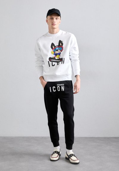 White sweatshirt with a cartoon graphic and "ICON" text, paired with black joggers featuring "DSQUARED2 ICON" branding. Black cap and two-tone shoes.