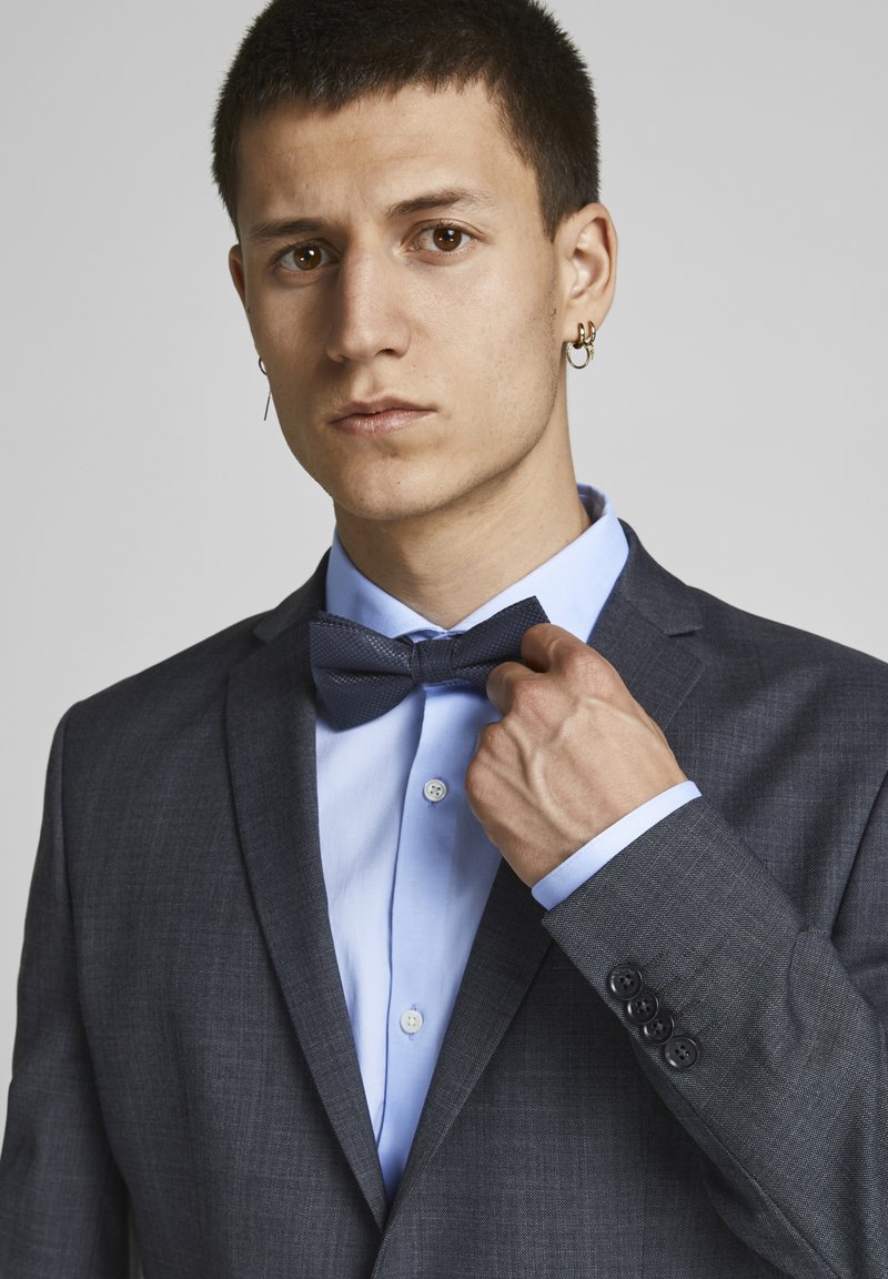 Chaqueta de traje gris con pajarita texturada azul oscuro sobre una camisa azul clara. Presenta puños abotonados y un corte ajustado. Apariencia pulcra y profesional.