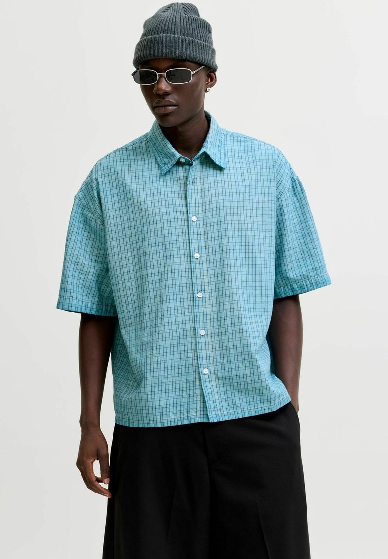 Homme portant un pantalon noir, une chemise bleu clair à manches courtes avec un motif à carreaux, un bonnet en maille gris et des lunettes de soleil ovales, debout devant un fond blanc.