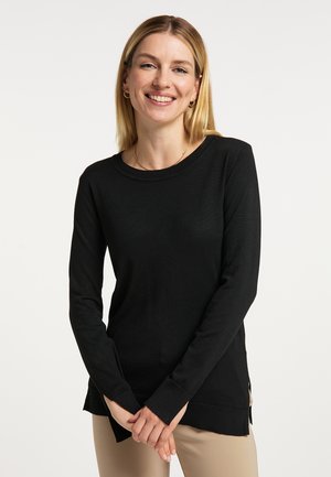 Lächelnde Frau mit schwarzem Langarmpullover und beigen Hosen, steht mit vor dem Körper verschränkten Händen vor einem einfarbigen Hintergrund.
