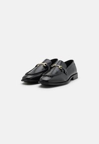 Mocassins en cuir noir avec un design cousu, des accents en métal doré et un petit talon, présentant une texture lisse et une silhouette classique.