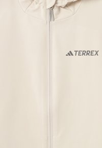 Giacca zip-up beige chiaro con cappuccio. Realizzata in tessuto morbido, presenta un logo TERREX grigio in alto a sinistra e una zip centrale.