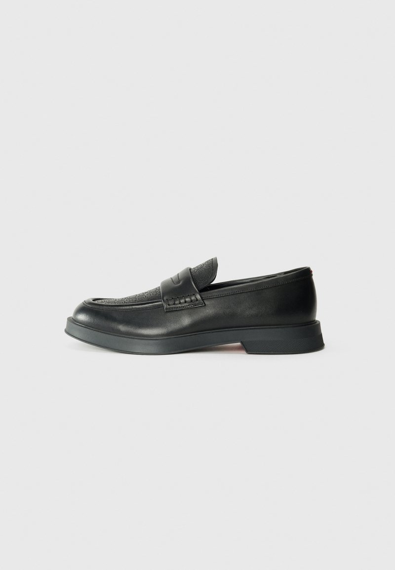 Mocasín de cuero negro con un upper texturizado, suela plana y acentos en relieve. Presenta un diseño sencillo y punta redondeada.