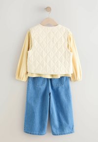 Gul langærmet skjorte lagdelt under en creme farvet quiltet vest, parret med blå denim shorts med to baglommer, hængt op på en træbøjle.