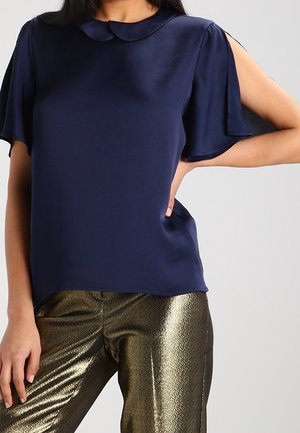 Femme portant un chemisier bleu marine à manches ouvertes aux épaules et un pantalon doré métallique, debout avec une main sur la hanche.