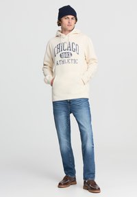 Felpa crema con grafica "Chicago 1983 Athletic" in blu navy, tasca a marsupio, indossata con jeans blu e scarpe marroni, e un berretto blu navy.
