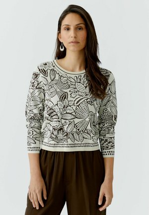 Vrouw met lang donker haar die een witte trui draagt met zwarte bloemen- en vogelpatronen, gecombineerd met donkerbruine broek.