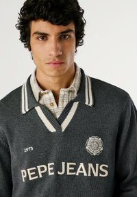 Siv sveter z belim prugastim ovratnikom, z napisom "PEPE JEANS" v belem in krožno logo; oblečen čez svetlo karirano srajco.