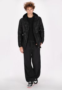 Sort camouflage zip-jakke med hætte, parret med løse sorte bukser og sporty sorte og hvide sneakers. Tekstureret stof, afslappet pasform.