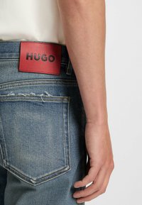 Siniset farkut, joissa on kuluneet taskudetailit ja punainen nahkapehmee, jossa lukee "HUGO" mustilla kirjaimilla. Käsi näkyvissä.