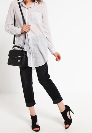Weißes Hemd mit Knöpfen und einer Brusttasche, kombiniert mit hochgerollten schwarzen Jeans und schwarzen Cut-Out-High Heels. Eine kleine schwarze Handtasche wird getragen.