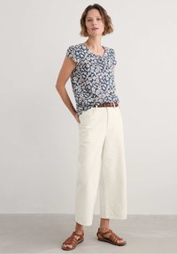 Halenka krátkého rukávu s květinovým vzorem v námořnické modři s bílými a oranžovými sedmikráskami; spárovaná s bílými kalhotami s širokými nohavicemi a hnědými sandály.