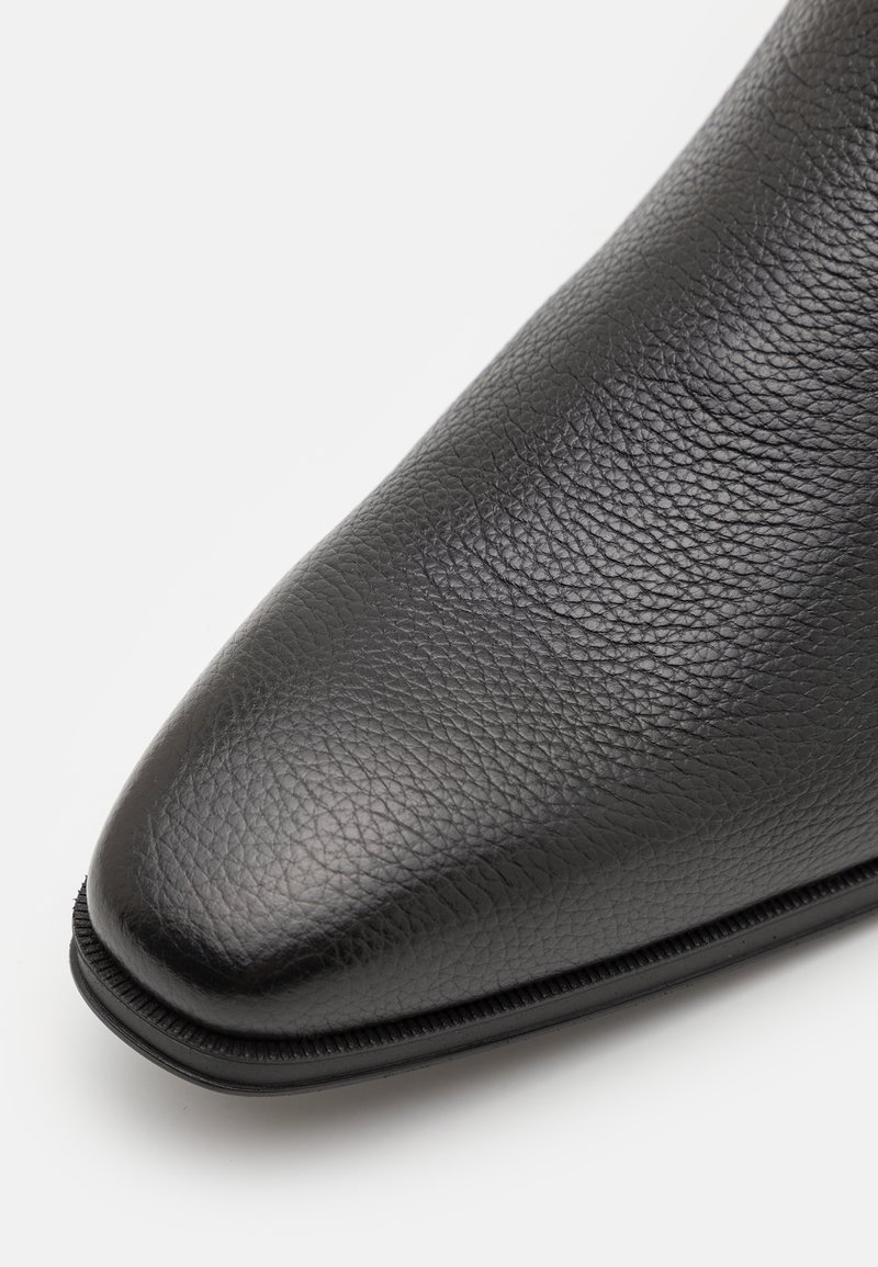 Botte de cheville en cuir noir avec une finition lisse et texturée, bout pointu et silhouette élancée. Présente un détail de couture subtil le long de la semelle.