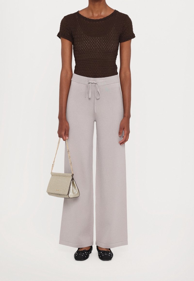 Personne portant un haut texturé marron foncé à manches courtes, un pantalon large gris clair avec cordon, des ballerines noires cloutées, tenant un petit sac à main beige.