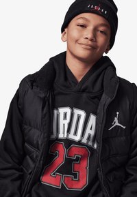 Colete acolchoado preto sobre um hoodie preto com um design vermelho e branco "Jordan 23". Um gorro preto com o logótipo "Jordan" completa o conjunto.