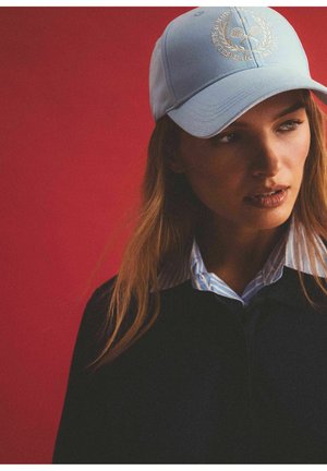 Jasnoniebieska czapka baseballowa z wyszywanym logo, noszona z ciemną bluzką i paskowanym kołnierzykiem, na modelu na tle czerwonym.