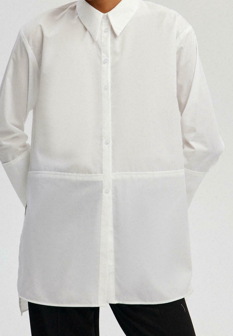 Personne portant une chemise blanche à manches longues avec col et pantalon noir, mains positionnées derrière le dos, fond clair uni.
