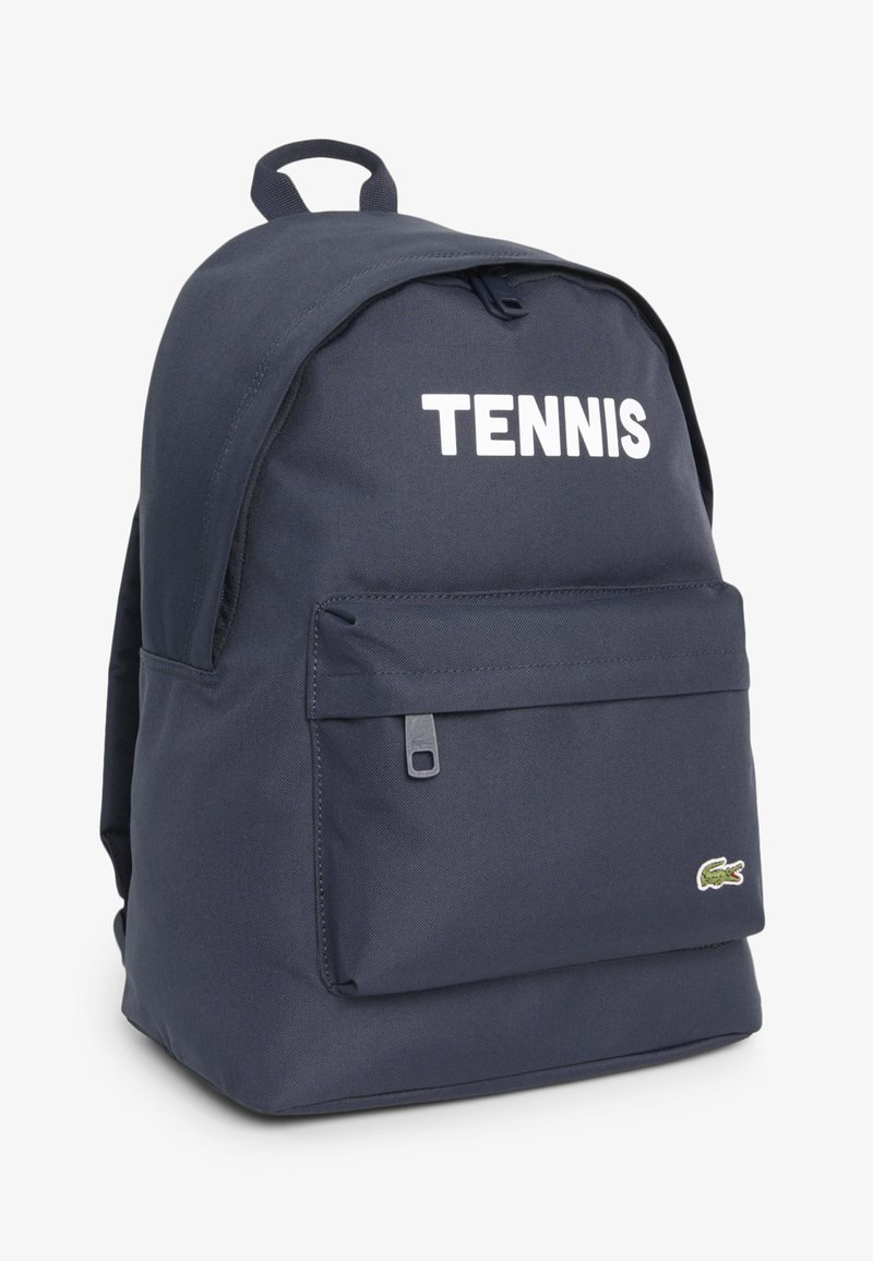 Zaino blu navy con tasca frontale con cerniera, scritta bianca "TENNIS" sulla parte superiore e piccolo logo verde coccodrillo nell'angolo inferiore.