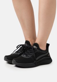 Black athletic shoes with a mesh upper, textured fabric, rubber sole, and lace-up design. Features a logo on the side and a cushioned collar.