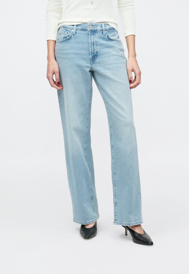 Ανοιχτό μπλε τζιν παντελόνι φαρδιάς γραμμής με ψηλή μέση, εμφανείς τσέπες στο μπροστινό μέρος και κούμπωμα με κουμπιά, σε συνδυασμό με μαύρες γόβες με μυτερή μύτη.