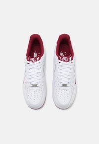 Tenisky Nike Air Force 1 s bielym koženým zvrškom, červenými textilnými akcentmi, perforovanou špičkou a bielymi šnúrkami. Metalické detaily na jazyku.