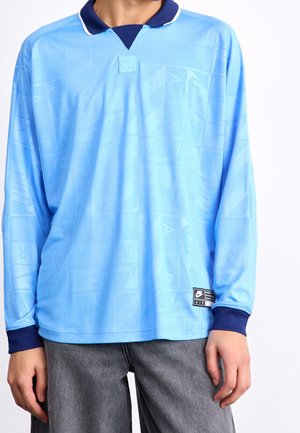 Polo Nike Air à manches longues bleu clair avec col et poignets bleu foncé, porté avec un pantalon gris, vue de face du cou jusqu'aux hanches.