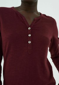 Burgundin pitkähihaisen paidan henley-tyylinen kaula-aukko, jossa on neljä hopeista nappia ja hieno tekstuuri. Kaksi sivusaumaa on näkyvissä.