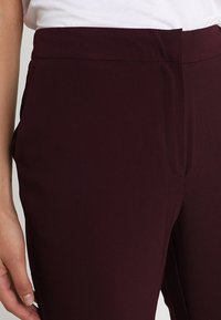 Burgundské kalhoty na míru s hladkou texturou, se zvýšeným pasem a bočními kapsami. Navrženy pro těsný střih.