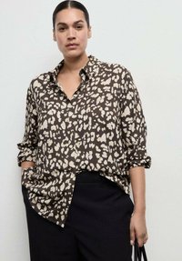 Rjava bluza z vzorcem leopardjega tiska v bež odtenkih. Ima ovratnik s prostimi gumbi in zavihane rokave. Nosijo se s črnimi hlačami.