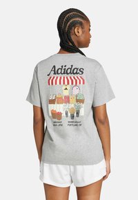 Grå kortärmad t-shirt med färggrafik av en marknad med frukter och grönsaker, texten "Adidas" och detalj av rödrandig markis på baksidan.
