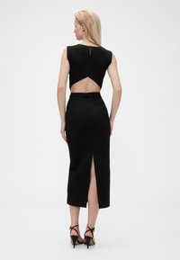 Melna kleita ar atvērumu mugurpusē, pieguļošs dizains, ceļgala garuma svārki, sānu šķēlums un gluda tekstūra. Valkāta ar melniem kurpēm ar siksniņu.