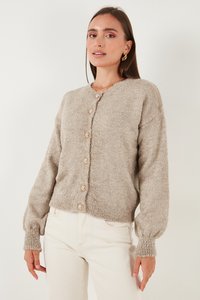 Žena oblečená v béžovém strukturovaném cardiganu s knoflíky a nabíranými rukávy a bílých kalhotách, stojící před jednoduchým světlým pozadím.