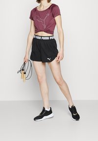 Bordeaux kortärmad t-shirt med en vit Puma-logotyp, tillsammans med svarta atletiska shorts som har en vit Puma midjeband. Håller i ett hopprep.