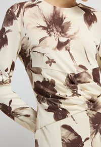 Długopisowa bluzka z miękkiego materiału w kremowej bazie z brązowymi kwiatowymi wzorami, o wyrafinowanej konstrukcji i detalach z marszczeniem w talii.