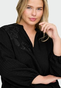 Blusa negra con tejido texturizado, que presenta mangas abullonadas y bordado floral en el hombro izquierdo. Diseño de escote en V.