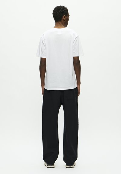 Weißes T-Shirt mit kurzen Ärmeln und rundem Ausschnitt, kombiniert mit einer lockeren schwarzen Hose, von hinten vor einem schlichten Hintergrund gezeigt.