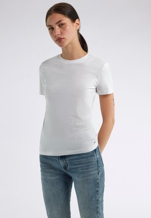 Donna con capelli scuri raccolti in una coda di cavallo indossa una semplice t-shirt bianca a maniche corte e jeans blu, in piedi davanti a uno sfondo grigio chiaro.