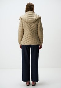 Beige gewatteerde pufferjack met een capuchon, voorzien van een chevronpatroon en elastische manchetten, gecombineerd met donkere wijde jeans.