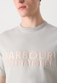 Světle šedé bavlněné tričko s žebrovaným kulatým výstřihem a embosovaným nápisem "BARBOUR INTERNATIONAL" v jemné béžové barvě.