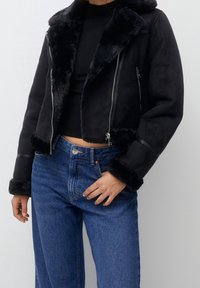 Chaqueta corta de falsa piel negra con cierre de cremallera, que presenta un amplio cuello y puños de piel, combinada con jeans de tiro alto azules.