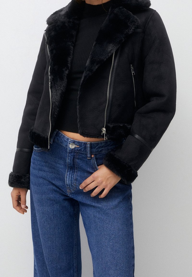 Chaqueta corta de falsa piel negra con cierre de cremallera, que presenta un amplio cuello y puños de piel, combinada con jeans de tiro alto azules.