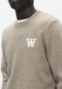 Beige sweater i blødt stof med rund hals og ribstrikkede ærmegab, med en hvid "W"-logo på brystet.