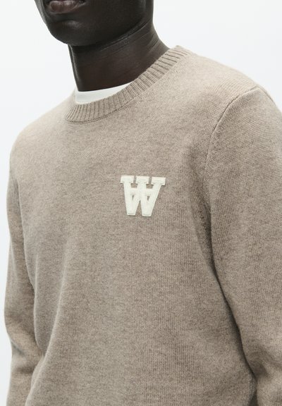 Pull beige en tissu doux avec un col rond et des poignets côtelés, présentant un logo blanc "W" sur la poitrine.
