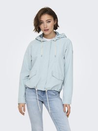 Femme portant une veste à capuche bleu clair avec cordons et un jean délavé clair, debout dans une posture décontractée devant un fond uni.