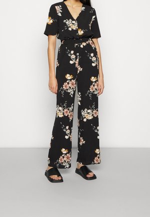 Musta kukallinen haalari, jossa on lyhyet hihat, V-pääntie, joustava vyötärö ja leveät lahkeet. Sisältää sinapinvärisiä ja vaaleanpunaisia kukkakuoseja. Yhdistetty mustien sandaalien kanssa.