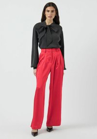 Haut noir à manches longues avec un grand nœud au col, associé à un pantalon rouge taille haute et large, et des talons noirs pointus.